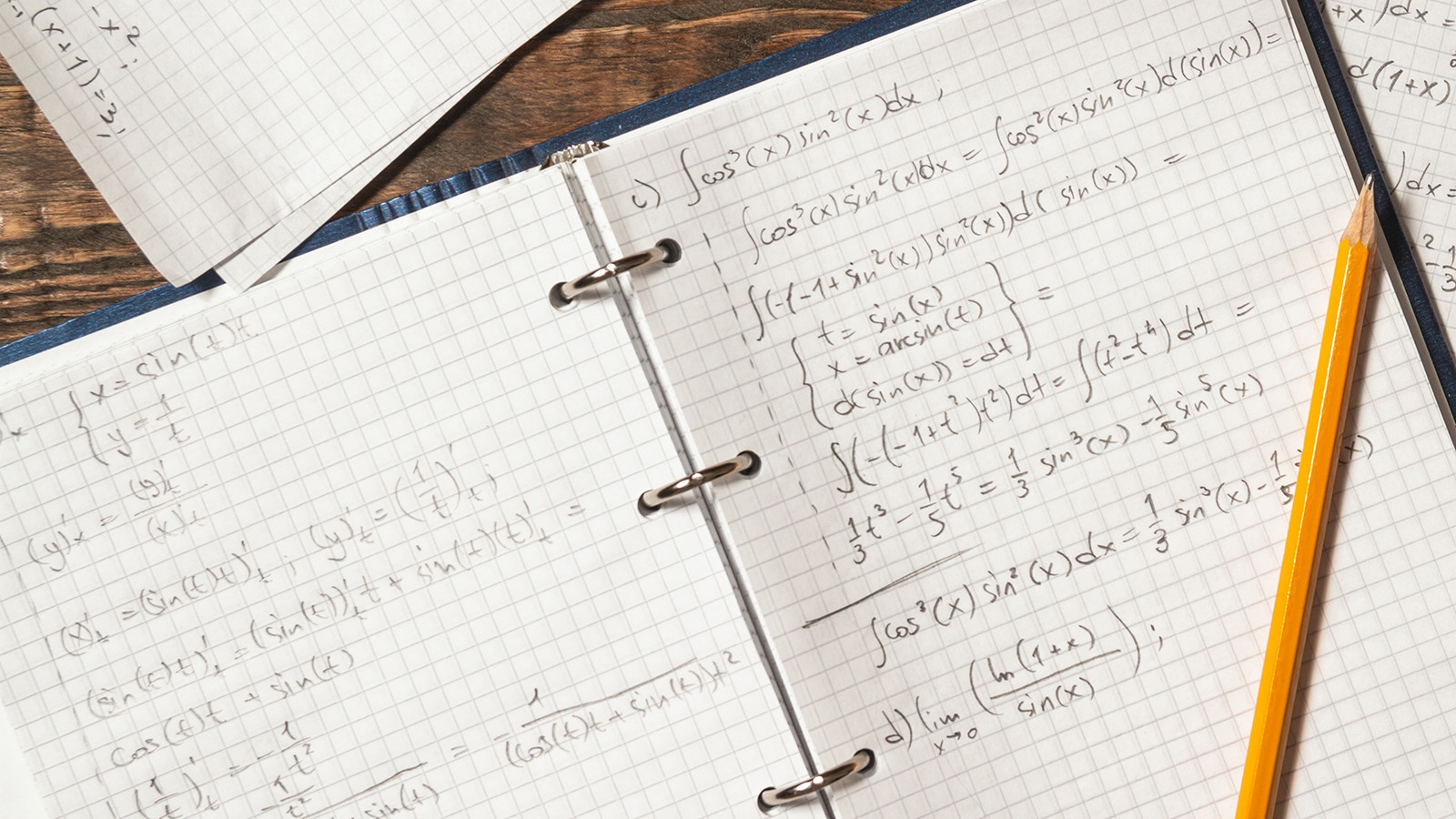 An open notebook showing calculus math problems, with a pencil laid across the top. Additional papers are strewn across the top of the image.